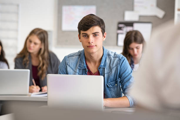 High school guy using laptop in classroom College student studying in library at the university with his classmates in background. High school young man using laptop for school work during computer lesson. Portrait of satisfied university guy sitting in classroom and looking at camera while working on laptop. 16 17 years stock pictures, royalty-free photos & images