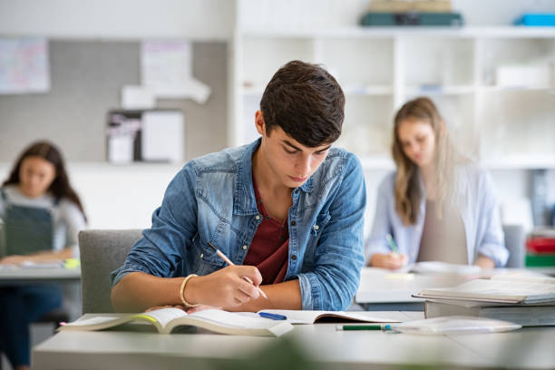 Teen student studying in classroom Focused young man taking notes from books for his study. College student sitting at desk with books for finding information in high school library. Guy studying in classroom and completing project with highschool classmates in background. exam student university writing stock pictures, royalty-free photos & images