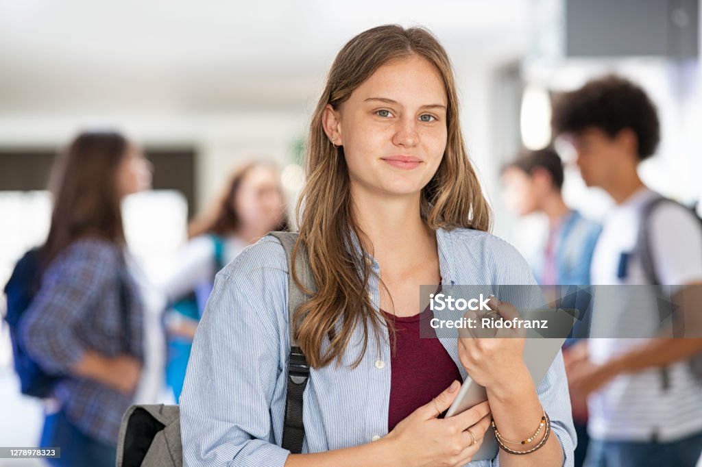 School girl holding digital tablet in college Portrait of happy satisfied girl standing in university hall with classmates in background. Pretty student holding digital tablet at high school and looking at camera. Smiling university young woman looking at camera after the end of the lessons. Teenager Stock Photo