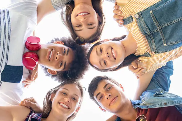 Close up face of happy multiethnic friends embracing in circle and looking down under blue sky. Group of smiling guys and beautiful girls bonding and having fun outdoors. Portraits of carefree best friends on a summer holiday looking at camera and laughing.
