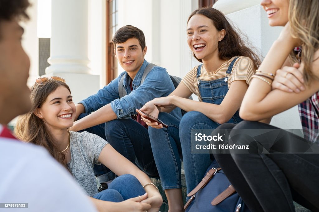 Teenager friends sitting together and laughing Group of happy young friends sitting in college campus and talking. Cheerful group of  smiling girls and guys feeling relaxed after university exam. Excited millenials laughing and having fun outdoor. Teenager Stock Photo