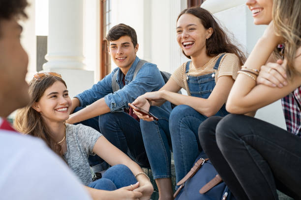 Teenager friends sitting together and laughing Group of happy young friends sitting in college campus and talking. Cheerful group of  smiling girls and guys feeling relaxed after university exam. Excited millenials laughing and having fun outdoor. group of students stock pictures, royalty-free photos & images