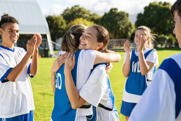 Smiling football players hugging on field after scoring a goal. Cheerful soccer teammates embracing while players clapping hands on victory. Successful girl soccer players embracing on field and celebrating after winning the match.