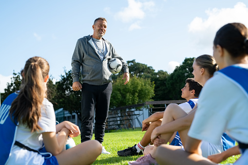 Football coach giving advices before the match. High school students listening to their sport teacher sitting on the soccer field. College professor give motivational speech to young football players.