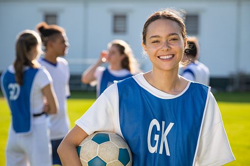 Cheerful soccer player holding a football and looking at camera. Portrait of young woman during training on soccer field. Satisfied high school student holding ball under the arm with her teammmats standing in background.