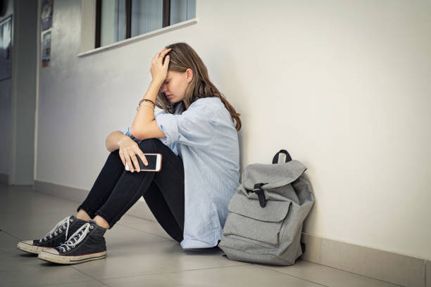 Cyber bullying at high school Upset and depressed girl holding smartphone sitting on college campus floor holding head. University sad student suffering from depression sitting on floor at high school. Lonely bullied teen in difficulty with copy space. online bullying stock pictures, royalty-free photos & images