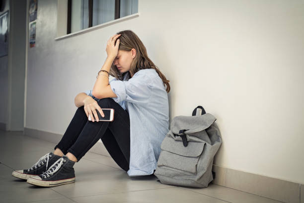 Cyber bullying at high school Upset and depressed girl holding smartphone sitting on college campus floor holding head. University sad student suffering from depression sitting on floor at high school. Lonely bullied teen in difficulty with copy space. grim stock pictures, royalty-free photos & images