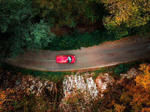 High angle image, taken by drone, depicting a topdown view of a red car driving on a country road through an autumn forest whose trees are beginning to taken on the yellow, orange and golden hues of fall. Room for copy space.