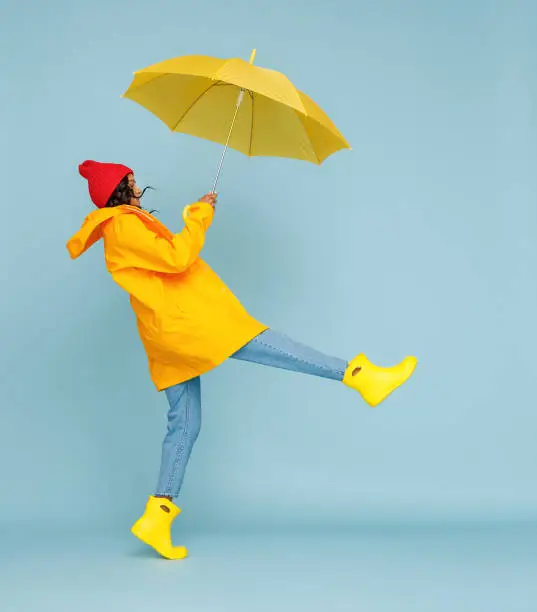 Side view of happy ethnic woman in outerwear and with umbrella leaping and having fun on rainy autumn day on blue background