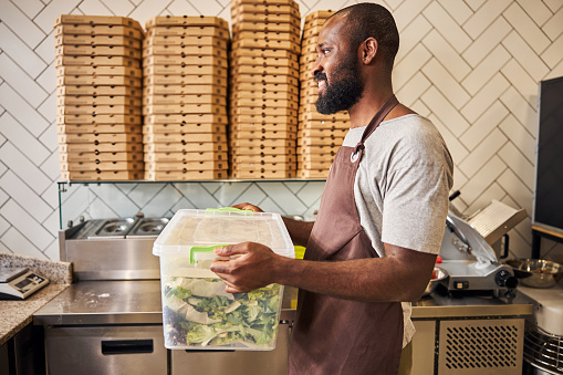 Handsome male worker in apron looking away and smiling while holding plastic box with fresh greens