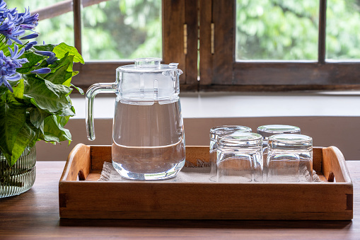A carafe of water and four empty glasses on a wooden tray next to the window, close up