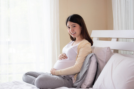 Young beautiful pregnant asian woman sitting on bed and touching belly near the window in bedroom. Pregnancy, motherhood, people and expectation concept.