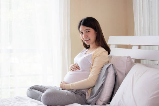 młoda piękna ciężarna azjatka siedząca na łóżku i dotykająca brzucha w pobliżu okna w sypialni. ciąża, macierzyństwo, ludzie i koncepcja oczekiwań. - window human pregnancy home interior women zdjęcia i obrazy z banku zdjęć