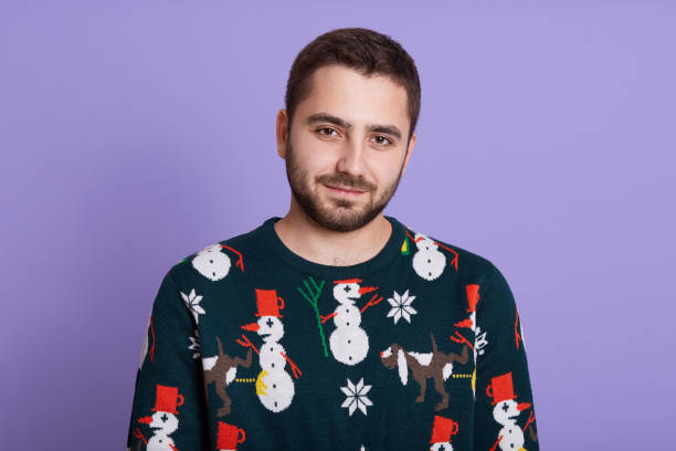 istock Bearded dark haired man wearing warm sweater with snowman looking at camera with calm expression, stands against lilac wall, looks directly at camera, 1279002784