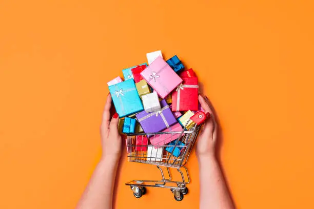 Christmas shopping cart full of colorful gifts, isolated on orange background. Top view with woman buying gifts. Online shopping for Christmas gifts.