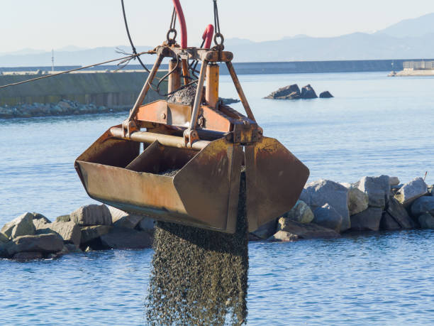 dredge clamshell bucket descargando grava en el agua de un puerto junto a la orilla para reponer una playa - draga fotografías e imágenes de stock