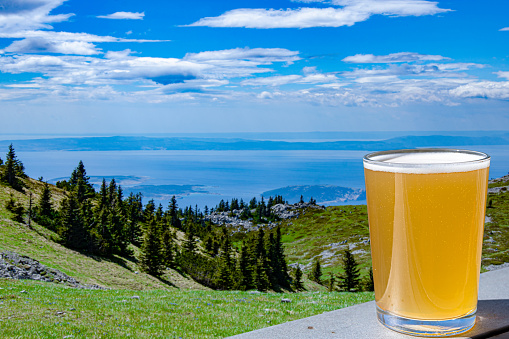 Glass of light wheat beer against mountains background. View of blue sea and green valley in the mountains.