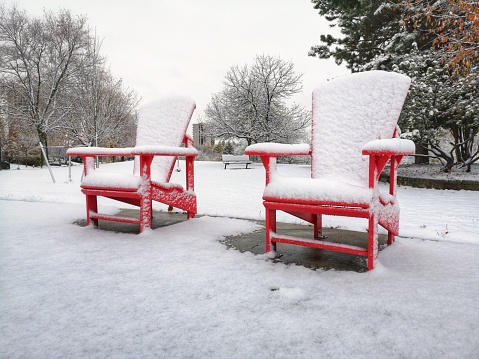 Two red wooden Adirondack Muskoka chairs covered with snow on winter day in park outdoor. Winter season Canadian landscape in Canada. Concept of forgotten lost lonely things on street outside.