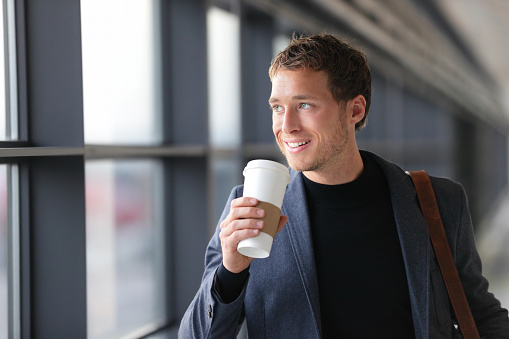 Businessman drinking coffee walking in airport. Casual urban professional smiling happy wearing suit jacket holding disposable coffee cup on travel. Handsome male model in his twenties.