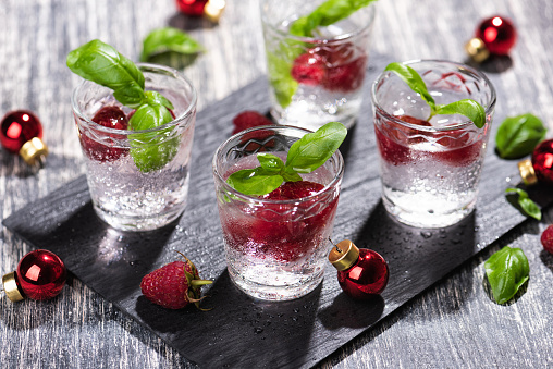 Alcohol shots of cocktail with a raspberry and basil on black stone tray. Glass of sparkling water and Christmas bubbles on wooden table