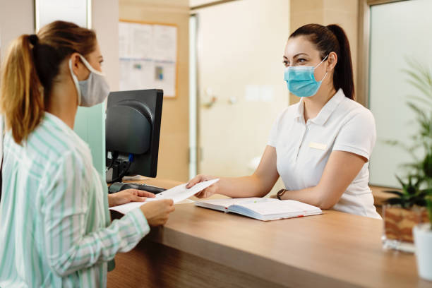 Health spa receptionist and her customer wearing face masks due to coronavirus pandemic. Female receptionist taking application form from her customer at health spa check in counter. They are wearing protective face masks due to COVID-19 pandemic. hotel reception stock pictures, royalty-free photos & images