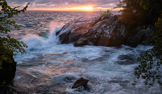 Big waves washing up on a cliff with sunset in the background and trees in the foreground
