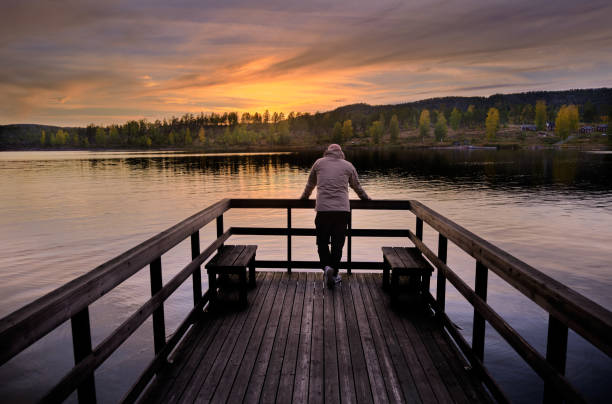 un hombre se para en un embarcadero y mira hacia el mar, la puesta de sol y el bosque en colores otoñales - norrland fotografías e imágenes de stock