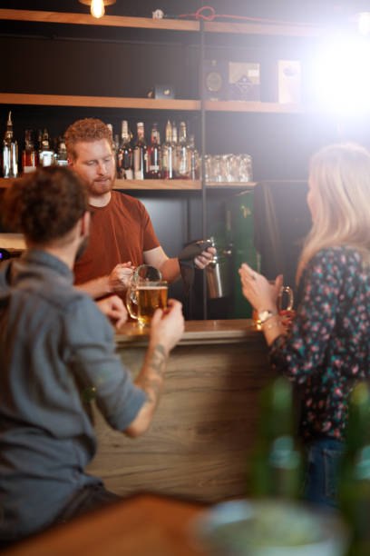 バーカウンターの隣に立つ若い魅力的なヒップスターカップル、ビールを飲み、バーテンダーとおしゃべり。バーテンダーはビールグラスを拭き、顧客と話しています。 - drinking couple bar counter bar ストックフォトと画像