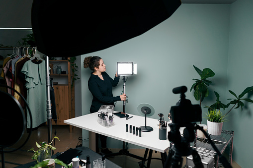 A young latin female vlogger standing and fixing the light of a recording set with beauty products and cosmetics.