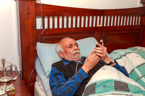 um indiano asiático de 72 anos visto se recuperando em sua cama em casa após sua cirurgia de vesícula biliar laparascópica; ele está muito ocupado com seu telefone inteligente. - medical oxygen equipment healthcare and medicine 70s mature adult - fotografias e filmes do acervo