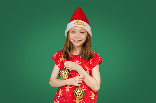 Happy Young Girl In Christmas Outfit Playing With Gold Christmas Baubles On Deep Green Background With Copy Space