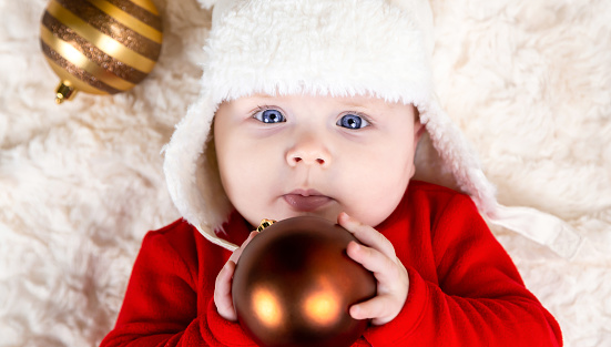 Cute toddler biting xmas ball, lying on the floor top view. A cute little girl in a red dress and white hat expresses emotions. Christmas concept with little kid