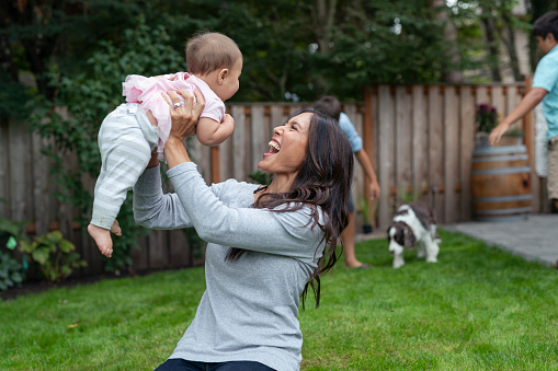 A beautiful mother of Pacific Islander descent lifts her baby girl above her head while spending a relaxing afternoon outside in the backyard. The woman's older kids play with the family dog in the background.