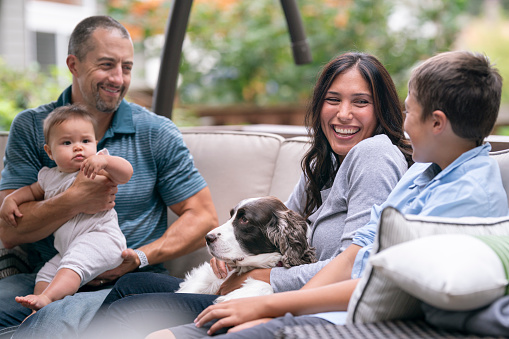 A mixed race family is relaxing together outside at home. A handsome man is affectionately holding his baby daughter while the family's dog lays on his wife's lap. The mother and father are laughing and looking at their elementary age son who has his back to the camear.