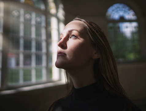 This image show a young caucasian woman looking up and through an antique window with her face being illuminated by sun rays.