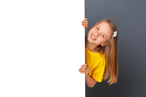 Funny teen girl in yellow clothes looks over a white wall / sign