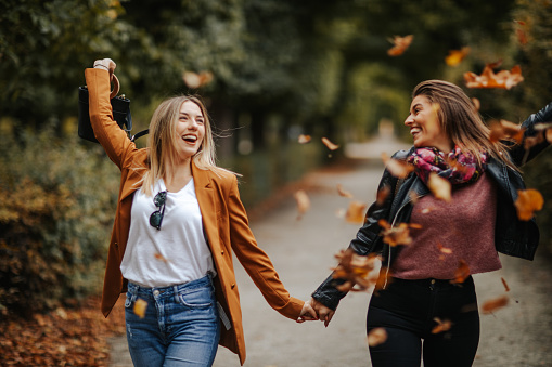 Happy young female friends holding hand and enjoying walk under falling leaves in autumn park on beautiful sunny day