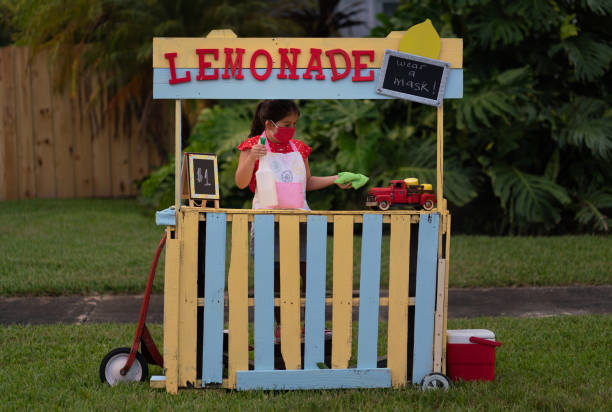 Outdoor rustic drink station with three glass beverage dispensers filled with citrus-infused lemonade and berries, set on wooden crates under a leafy garland against a stone wall, with glass tumblers ready for serving