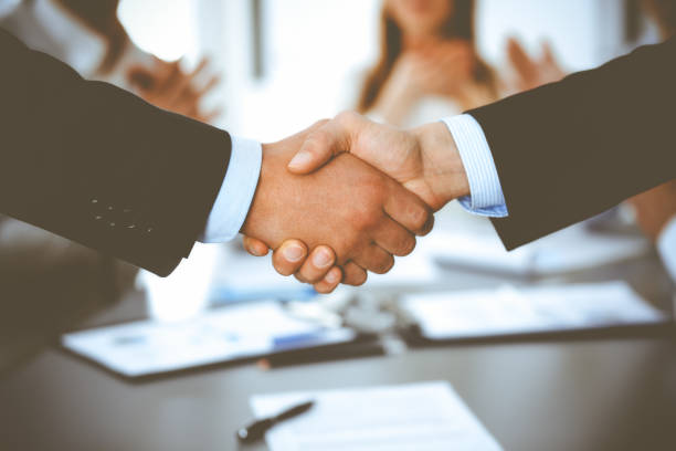 istock Business people shaking hands at meeting while theirs colleagues clapping and applauding. Group of unknown businessmen and women in modern white office. Success teamwork, partnership and handshake concept 1279101822
