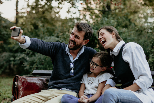Beautiful family sitting on the wooden bench, where mom and girl waited for the father to come after work so they can spend some joyful time at the park. Dad is taking a selfie with them with a big smile and excitement on his face.