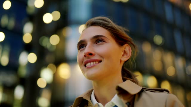 Panning closeup shot of confident young woman in trench coat standing in city street surrounded by bokeh lights and looking away dreamily with happy smile on face, her hair waving in wind