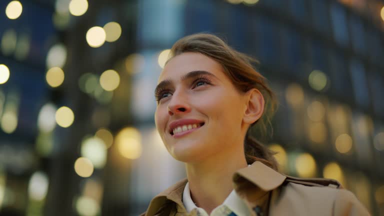 Panning closeup shot of confident young woman in trench coat standing in city street surrounded by bokeh lights and looking away dreamily with happy smile on face, her hair waving in wind