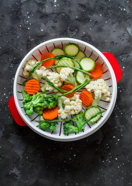 Photo of Cooking healthy food concept. Fresh vegetables broccoli, zucchini, carrots, asparagus in a steamer on a dark background, top view