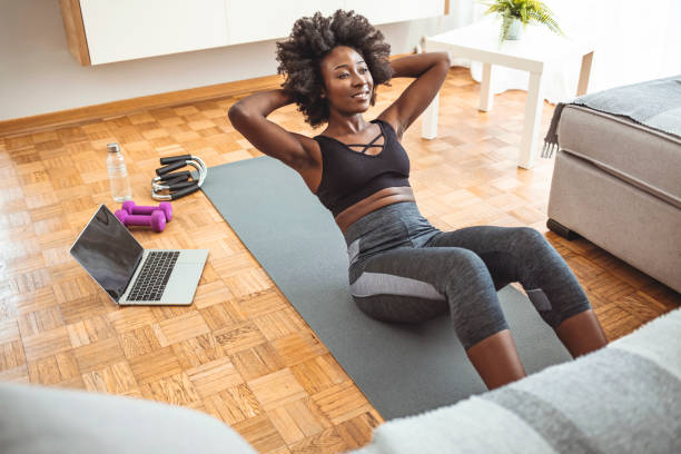 Young smiling athletic woman doing sit-ups at home. African american sportswoman doing abs on yoga mat near laptop in living room. African-American woman having an online training at home. Exercising on the floor sit ups stock pictures, royalty-free photos & images