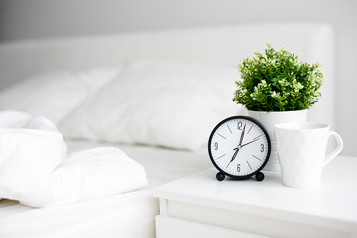 good morning concept - alarm clock and cup of coffee on bedside table and unmade bed at home or hotel