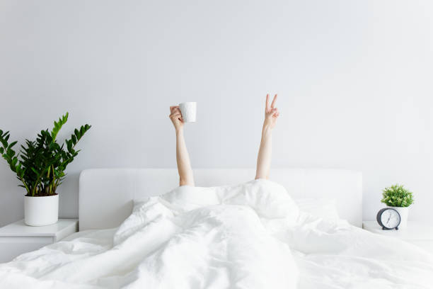 goedemorgenconcept - vrouwelijke handen met koffiekop en overwinningsteken die uit de deken thuis of hotel steken - bed stockfoto's en -beelden