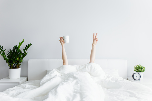 good morning and relaxation concept - female hands with coffee cup and victory sign sticking out from the blanket at home or hotel