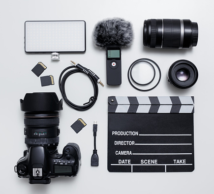 videography and photography equipment - top view flat lay of modern dslr camera, lenses, filters, microphone with windscreen, led light, memory cards and clapper board over white table background