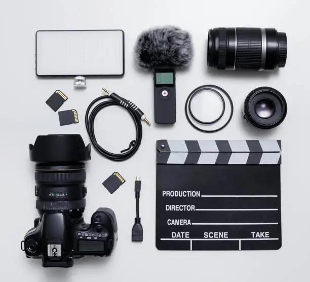 Photo of videography and photography equipment - top view flat lay of modern dslr camera, lenses, filters, microphone with windscreen, led light, memory cards and clapper board over white table
