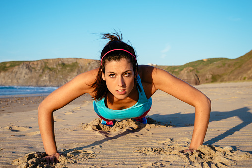 Fitness woman doing push ups at the beach on summer. Brunette sporty girl training hard.